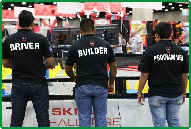 3 collage age students stand in front of a competition field with their backs to the camera. their shirts read: VEX U Driver, builder, and programmer 3 collage age students stand in front of a competition field with their backs to the camera. their shirts read: VEX U Driver, builder, and programmer